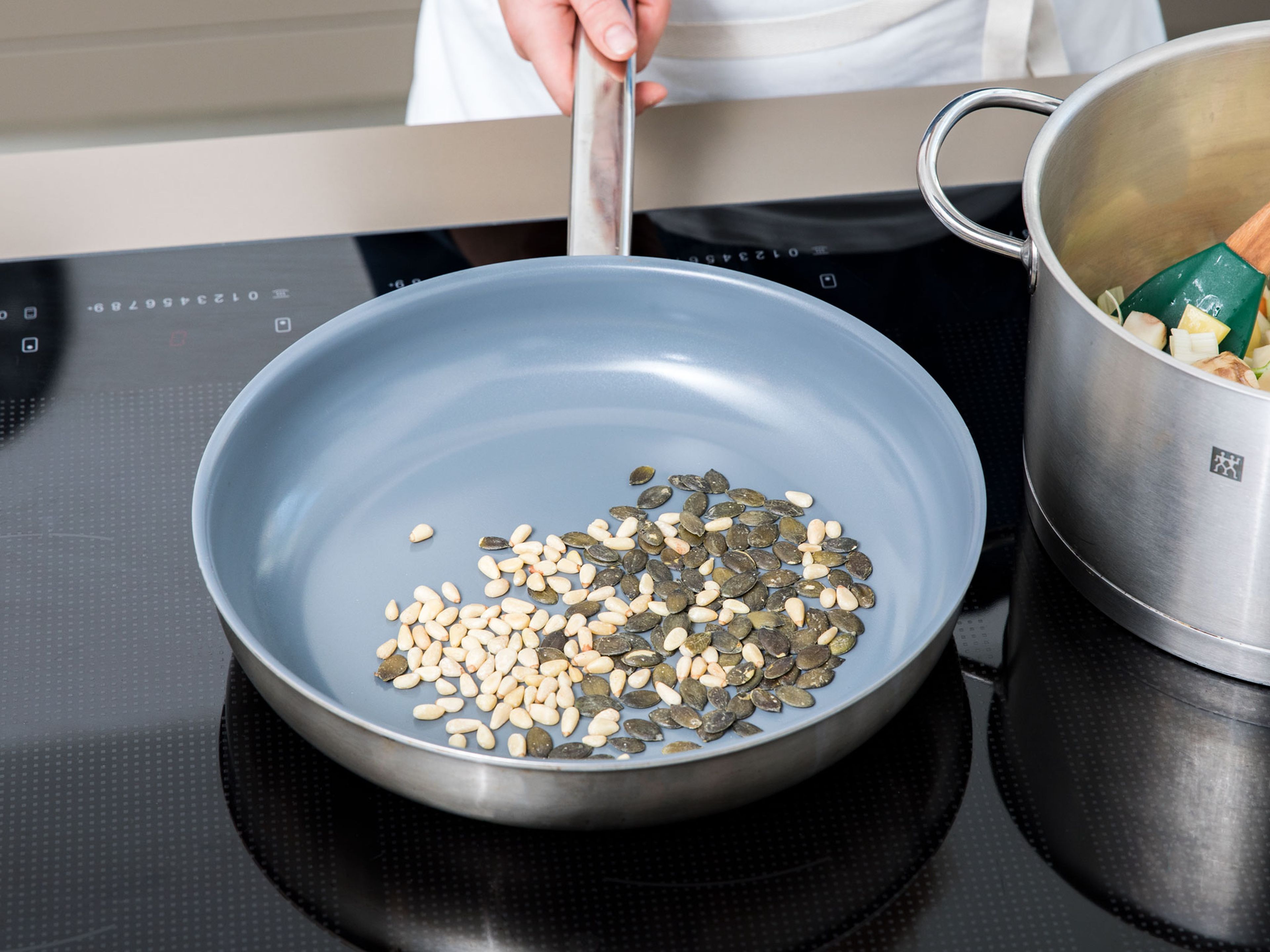 In der Zwischenzeit Pinienkerne und Kürbiskerne in einer fettfreien Pfanne anrösten, bis sie gebräunt sind und ihr Aroma entfalten. Die Kerne aus der Pfanne nehmen und beiseitestellen.