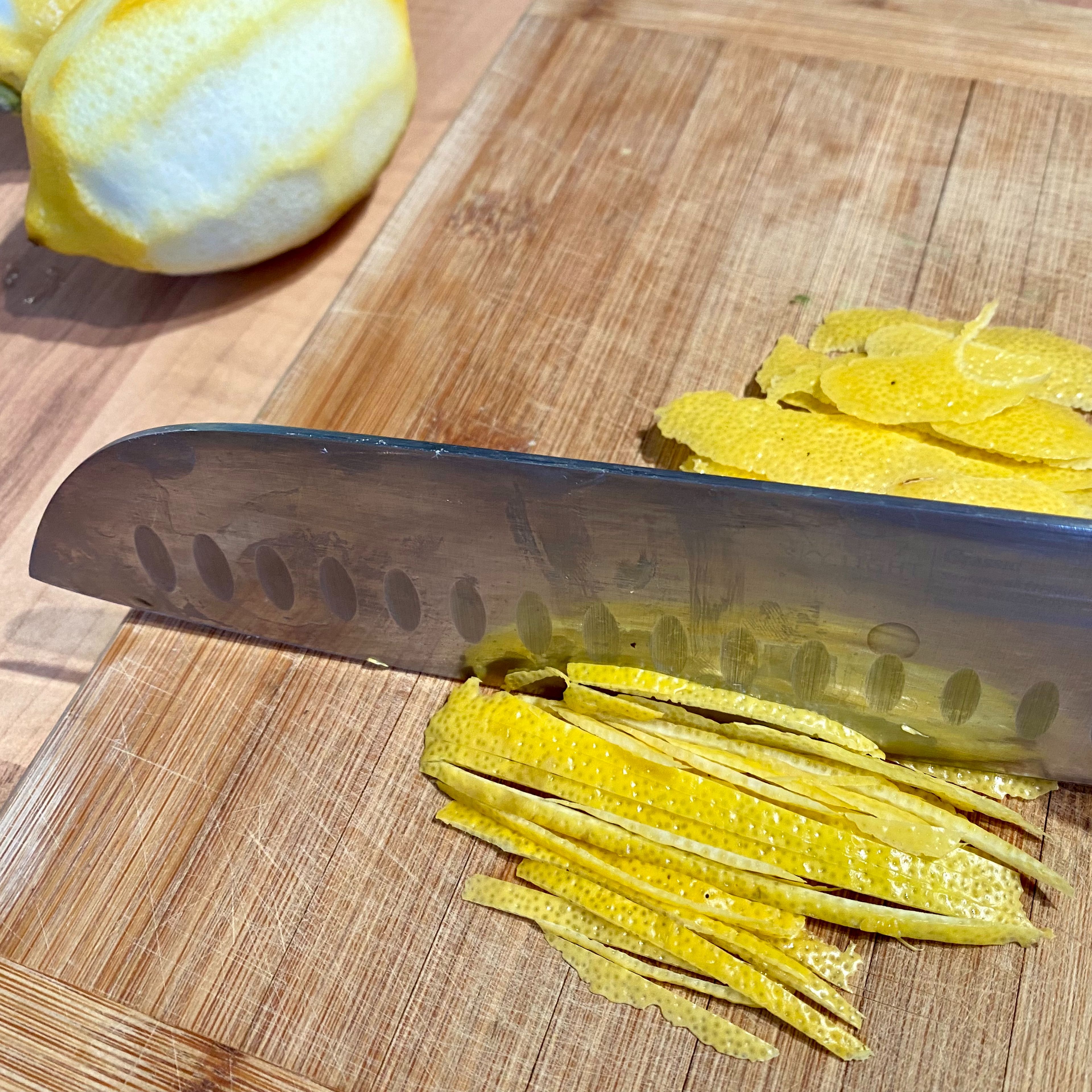 Währenddessen die Schale der Zitronen mit einem Sparschäler von einer Seite zur anderen abschälen und längs mit einem Messer in feine Streifen schneiden. Den Saft der beiden Zitronen in den köchelnden Sirup auspressen. Die geschnittene Zitronenzeste ebenfalls hineingeben und das ganze 10 Minuten köcheln lassen, danach beiseitestellen und abkühlen lassen.