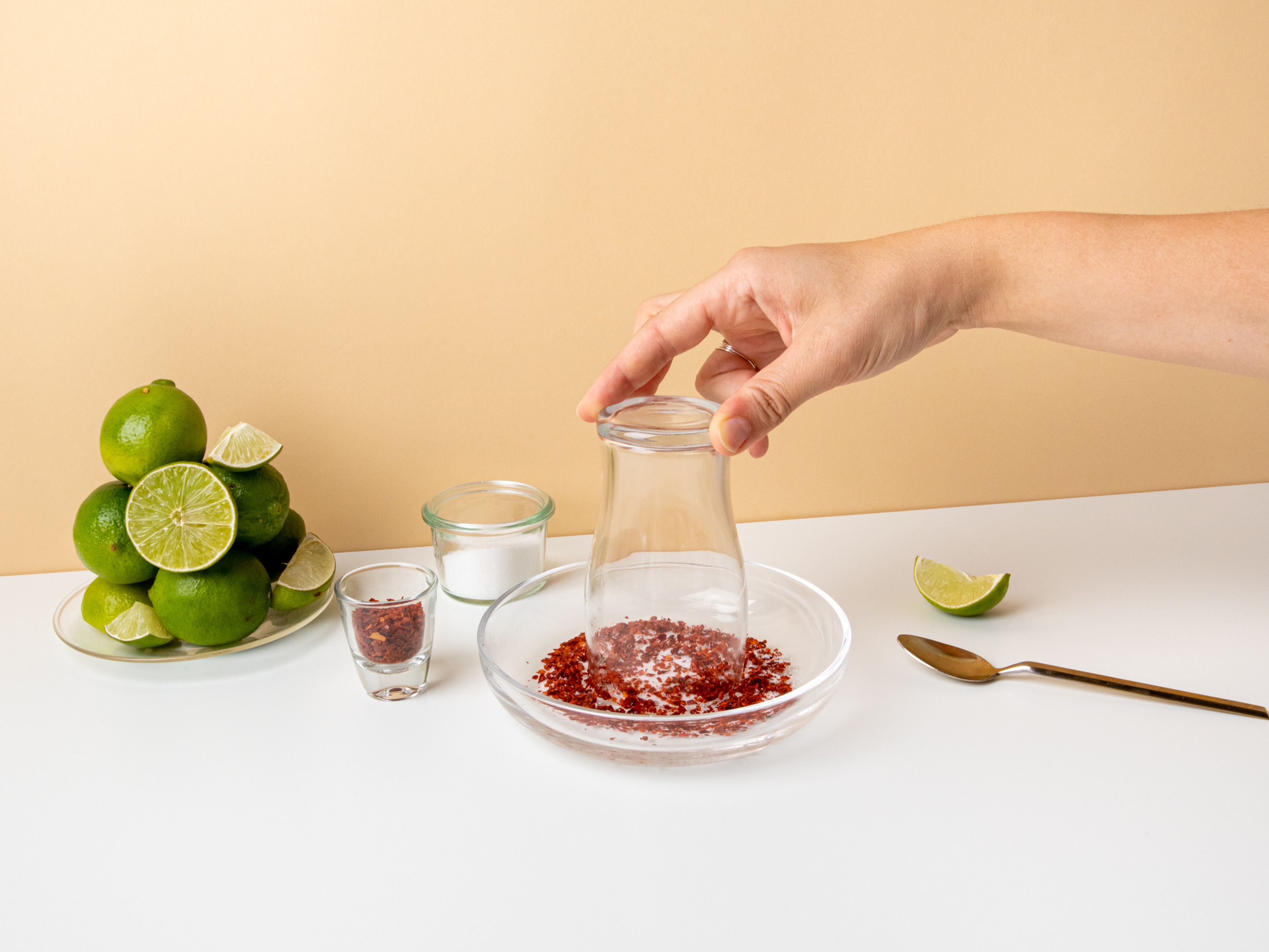 Für die Michelada die Chiliflocken, Zucker und grobes Meersalz auf einen Teller geben und vermengen. Eine der Limetten halbieren und das Servierglas mit einer Limettenhälfte einreiben und anfeuchten. Das Glas mit dem Rand nach unten in die Chili-Salz-Mischung tunken und so einen salzig-scharfen Rand erzeugen.