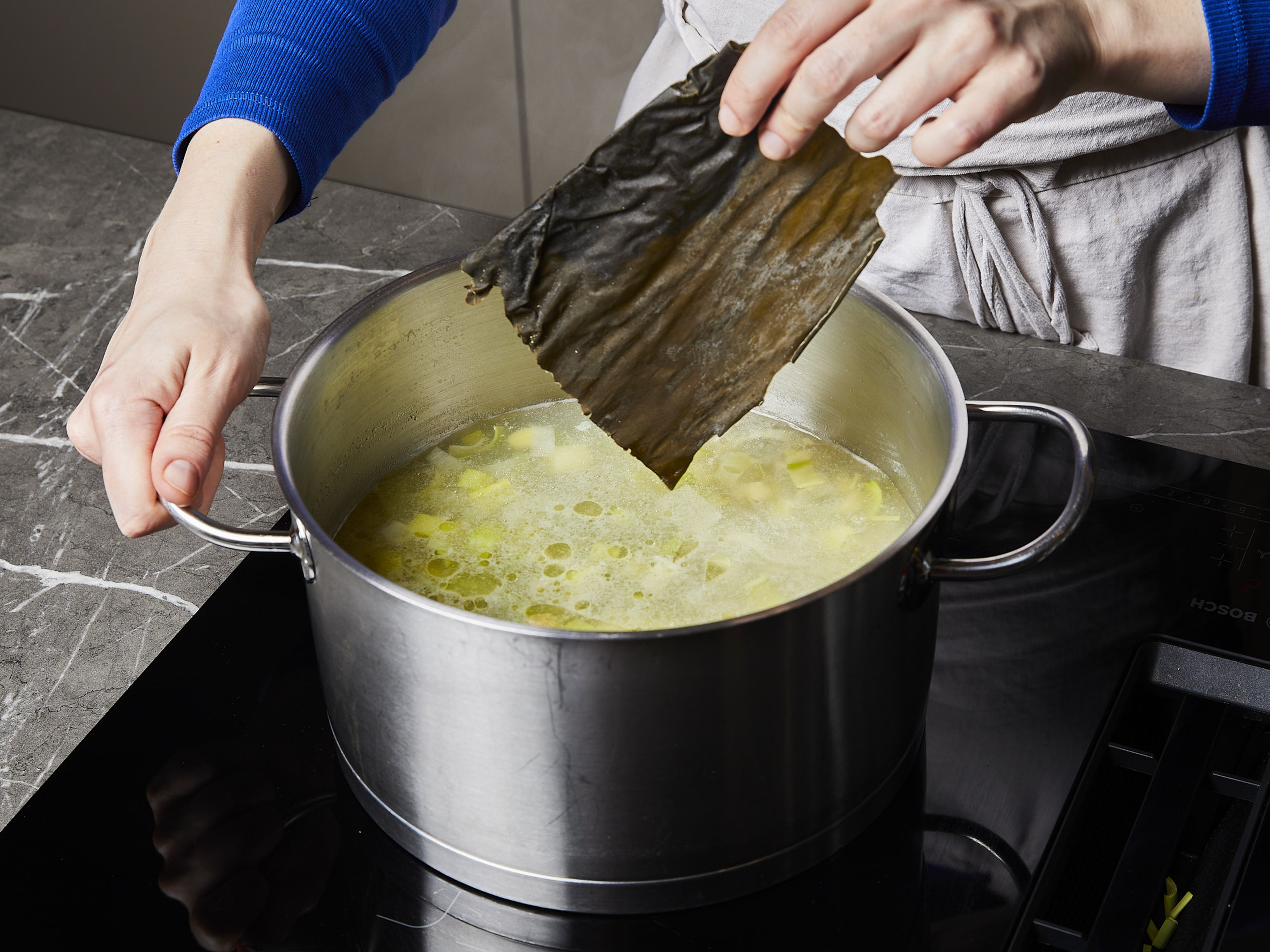 Für die Ramen, ⅚ des Knoblauchs und ⅔ des Ingwers grob hacken. Lauch der Länge nach halbieren und in Scheiben schneiden. Hühnerbrühe, Lauch, gehackten Ingwer und Knoblauch sowie die entbeinten Hähnchenschenkel in einen großen Topf geben und zum Kochen bringen. Ca. 15 Min. kochen lassen, dann das Fleisch aus dem Topf nehmen, etwas abkühlen lassen und beiseitestellen. Den Kombu in die Brühe geben und weitere 15 Min. kochen, dann alles durch ein Sieb abgießen.