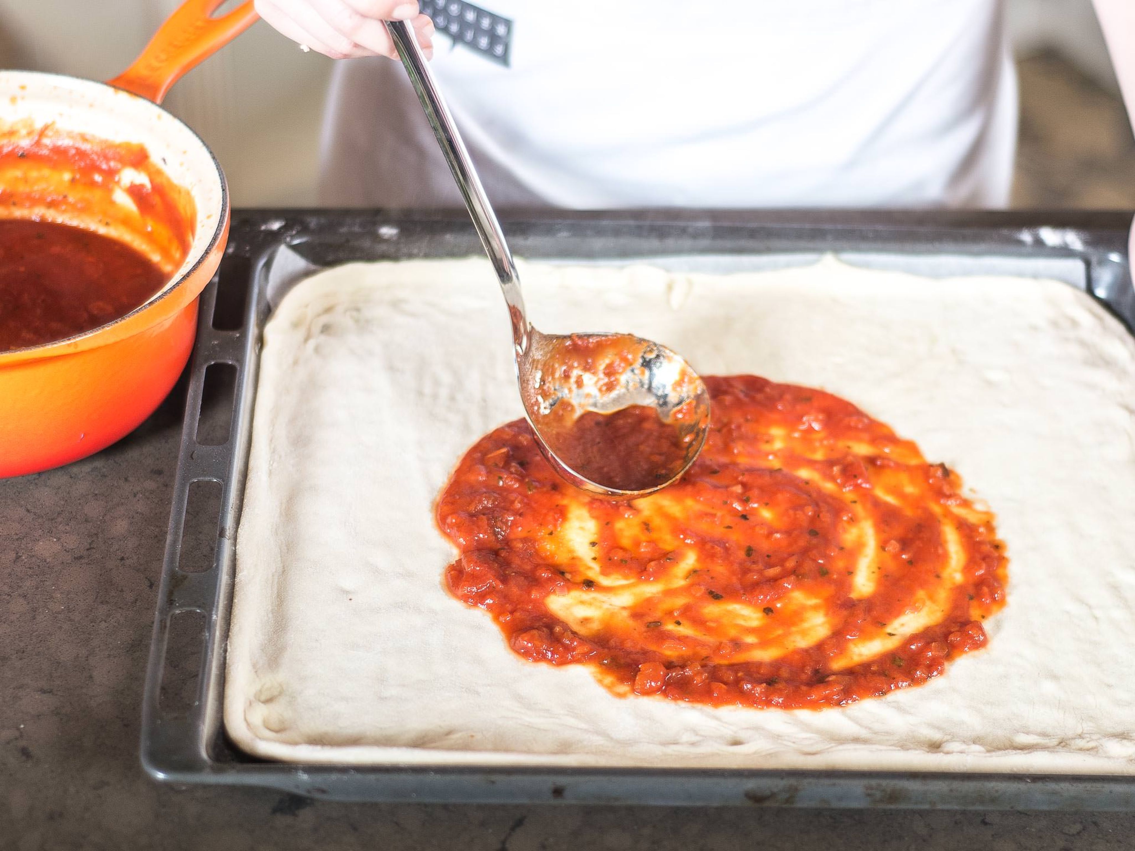 Die Tomatensoße mit einer Schöpfkelle gleichmäßig auf dem Teig verteilen.